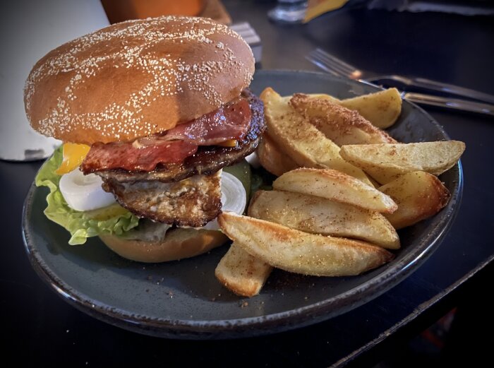 Hamburgare med bacon, ost och grönsaker, serverad med steak house fries på en mörk tallrik.