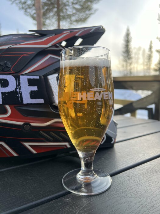 Ölglas på ett bord med suddig bakgrund av en hjälm och träd, utomhusmiljö.