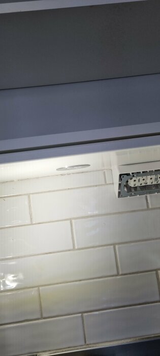 Vita kakelplattor på vägg med öppen eluttagsbox och delvis synlig ledning under köksskåp.