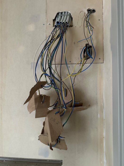 Oorganiserade elektriska kablar och en ihopklippt bit kartong på en gipsskiva under renovering.