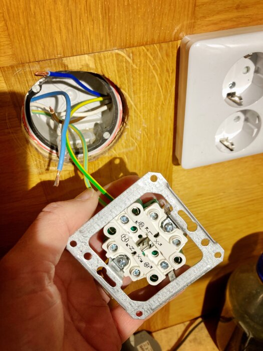 Hand håller en eldosa med kablar redo för installation bredvid ett befintligt vägguttag på en träpanelvägg.