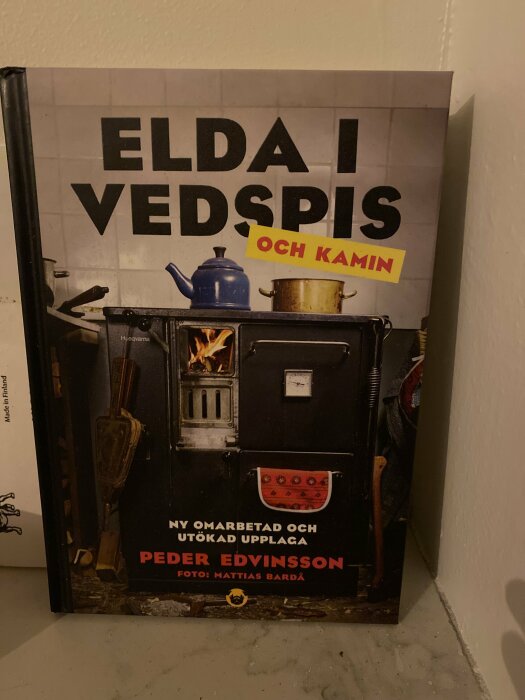 Bok med titeln "ELDA I VEDSPIS och KAMIN" av Peder Edvinsson, bild på vedspis med tekanna.