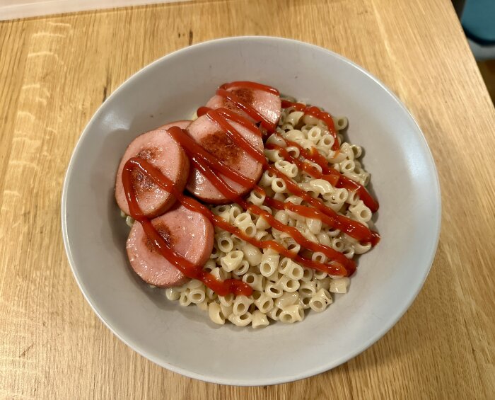 En tallrik med kokt makaroni och skivad stekt korv toppad med ketchup.