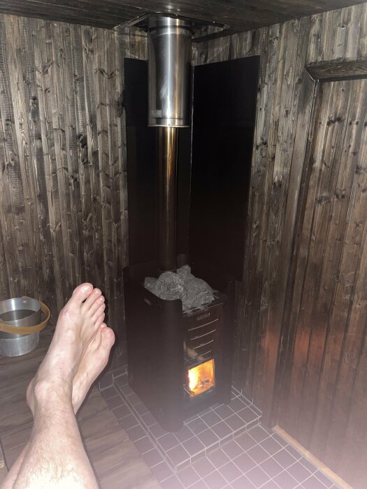 Interiör av bastu med tänd vedeldad bastuaggregat och person som sträcker ut benen.