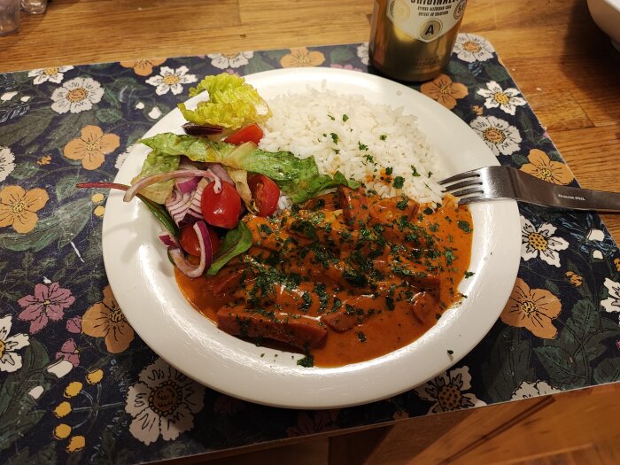 En tallrik med korvstroganoff, ris och sallad på ett blommigt duk.