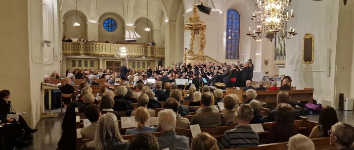Kör och orkester framför musik för publik i kyrka med ljuskronor och altare.