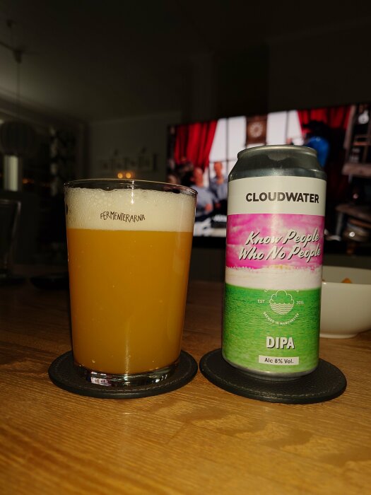 Grumlig, humlig öl i glas bredvid Cloudwater ölburk på ett bord med suddig TV i bakgrunden.