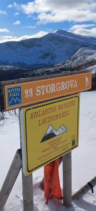 Skyltar för STORGROVA pist och lavinvarning i fjällterräng med snö och berg i bakgrunden.