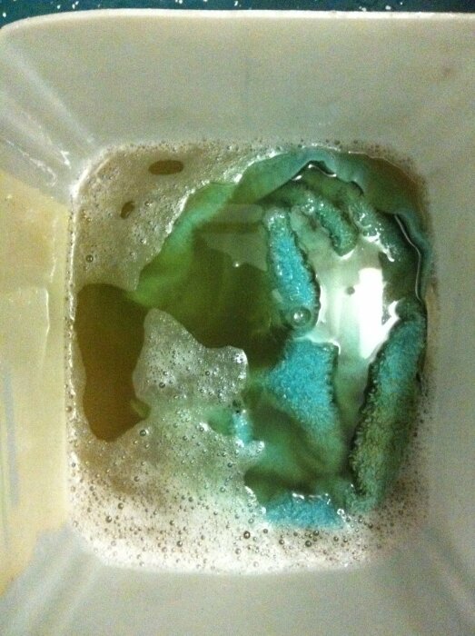 En sprayflaska och mikrofiberduk i en vask med en grön ammoniakblandning och skum.