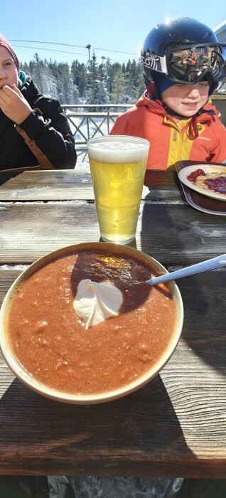 Skidåkare som äter lunch ute med tomatsoppa och öl på ett bord, med snötäckt landskap i bakgrunden.