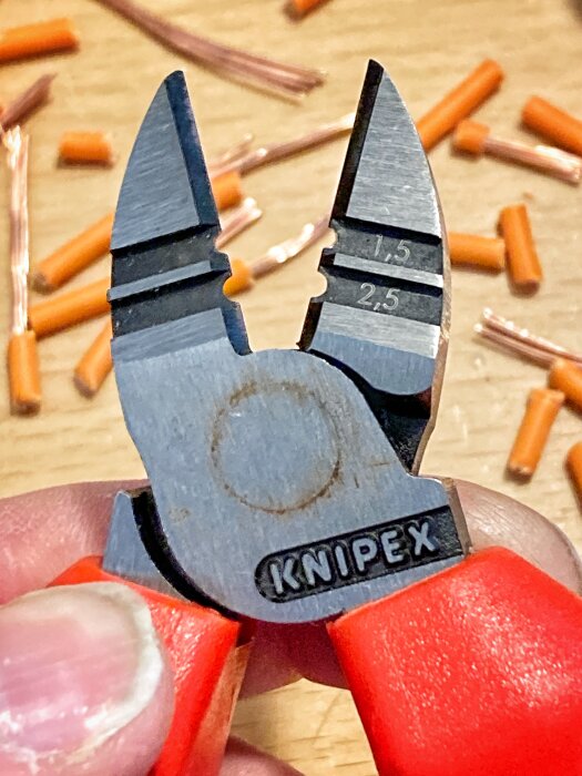 Närbild på en Knipex skaltång i hand, med kabelskalningsmärkningar och avklippta kabeländar i bakgrunden.