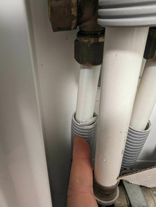 Finger som pekar på ett grått plaströr som läcker vatten vid anslutning till vit avloppsrör bakom metallventiler.