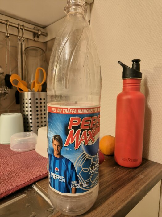 Gammal Pepsi Max-flaska med reklam med fotbollsspelaren på etiketten, på en köksbänk bredvid en röd vattenflaska och en apelsin.