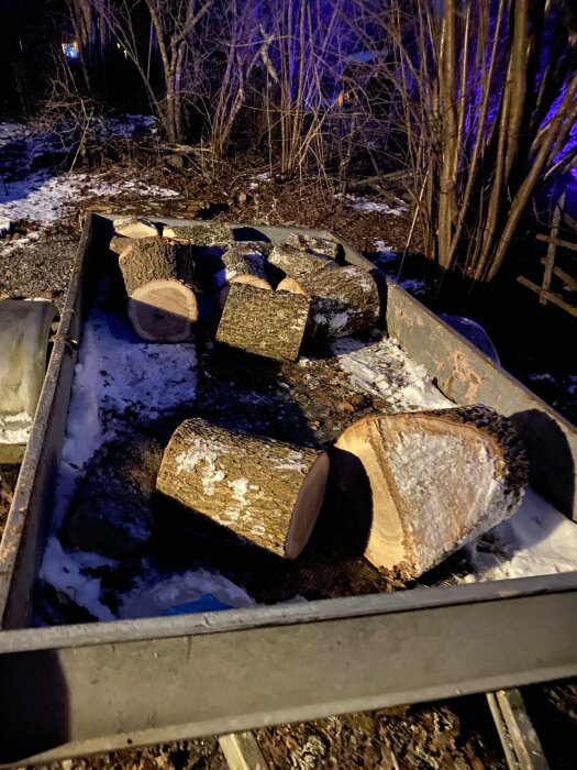 Vedstockar i en skottkärra på snötäckt mark under kvällsbelysning.