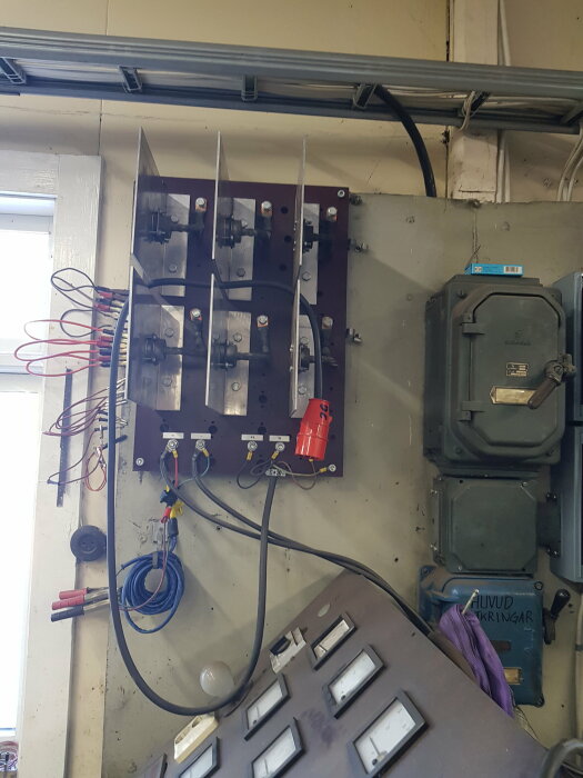 Trefasig likriktare med öppna komponenter och kablar i en verkstadsmiljö, bredvid säkringsskåp och strömbrytare.