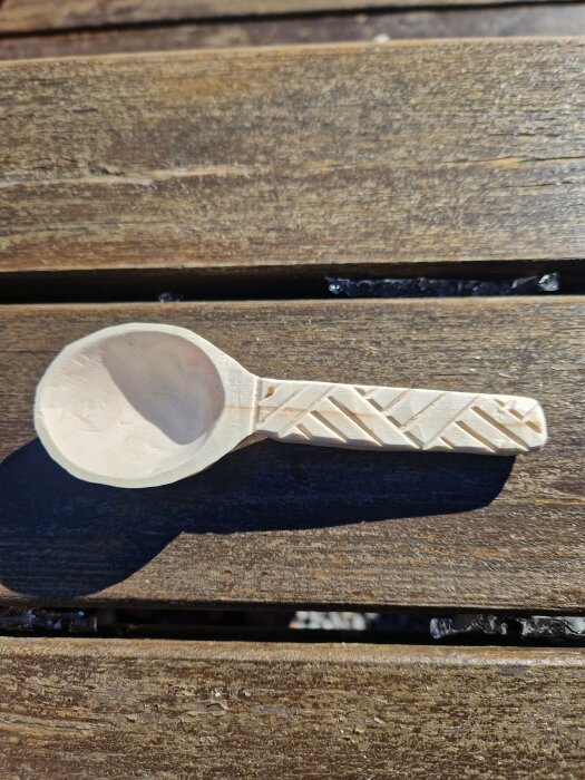 Handtäljd liten sked i lönn med mönster på skaftet, placerad på en träbänk.