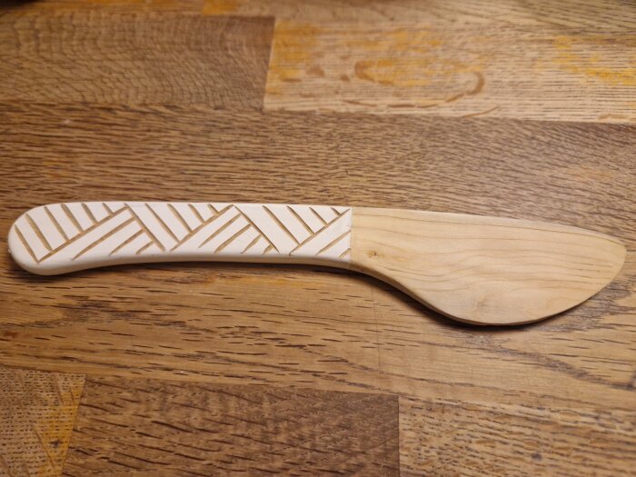Handtäljd smörkniv i lönn med dekorativt mönster på skaftet, ligger på träbord.