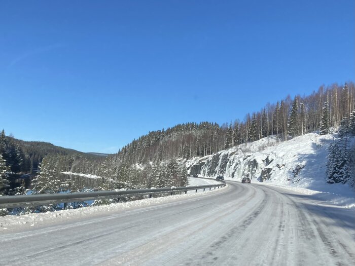 Vinterväg kantad av snötäckta träd och berg under en klarblå himmel.