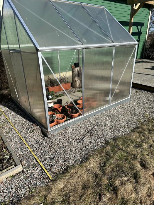 Växthus av aluminium och plast på grusunderlag med krukplanteringar och trädgårdsredskap inuti.