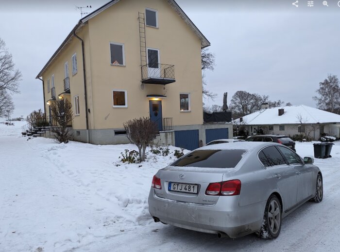 Gul tvåvåningshus med garage och snötäckt parkering, bil framför huset.