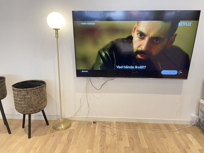 Väggmonterad TV med synliga sladdar och modem, strömkontakter synliga till höger, modern stående lampa till vänster.