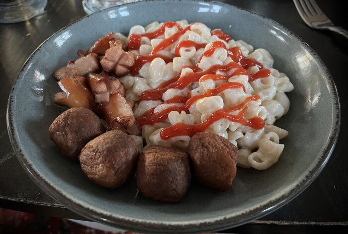 Måltid med stuvade makaroner, kycklingköttbullar, prinskorv och ketchup på en grå tallrik.