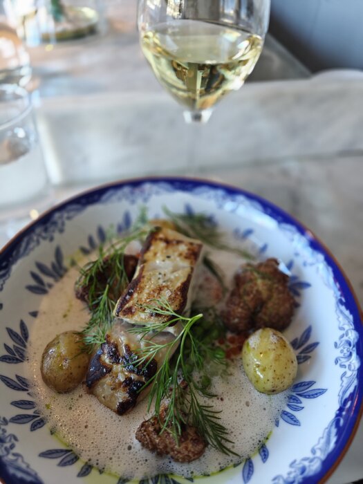 Tallrik med stekt sej, små potatisar och dill, i förgrunden med ett glas vitt vin suddigt i bakgrunden.