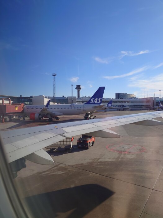 Vy från flygplansfönster över en flygplats med SAS-flygplan, servicefordon och torn i bakgrunden.