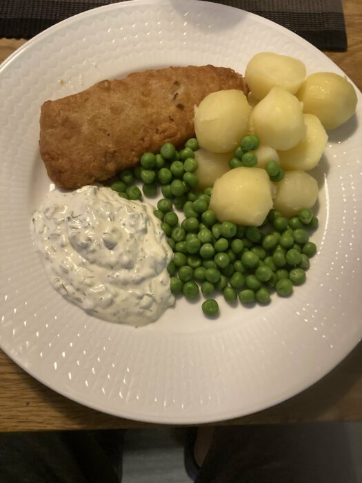 Panerad fisk med kokt potatis, gröna ärtor och dillsås på en vit tallrik.