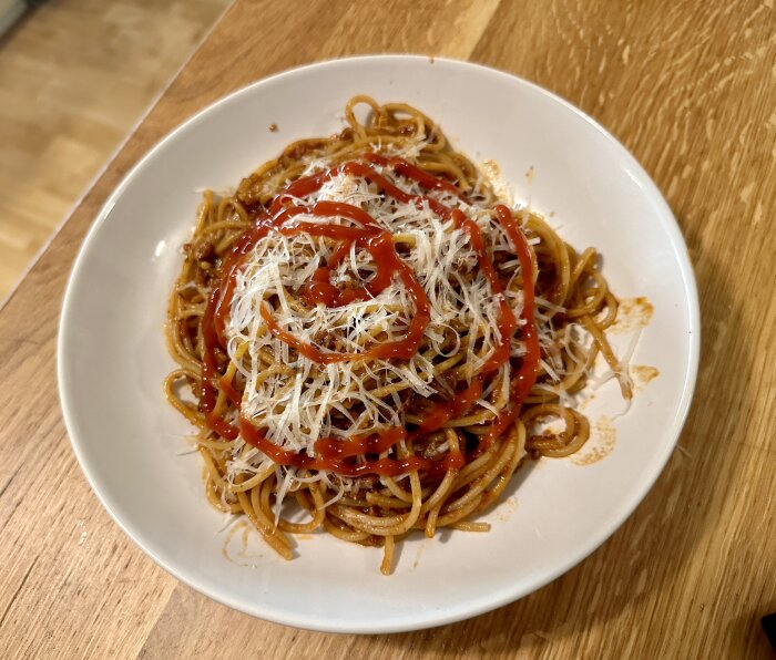 En tallrik spaghetti med köttsås, riven ost och ketchup på träbord.