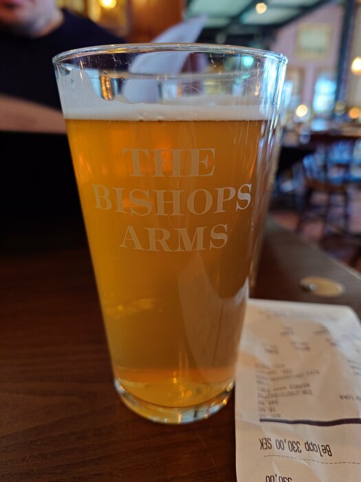 Ett ölglas fyllt med IPA märkt "THE BISHOP'S ARMS" framför en suddig barbakgrund.