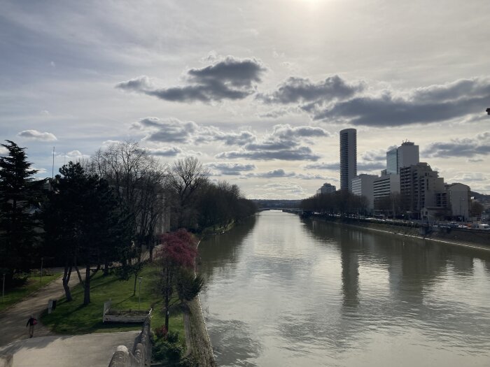 Stadsutsikt över en flod med promenadvägar, träd, och moderna byggnader under molnig himmel.