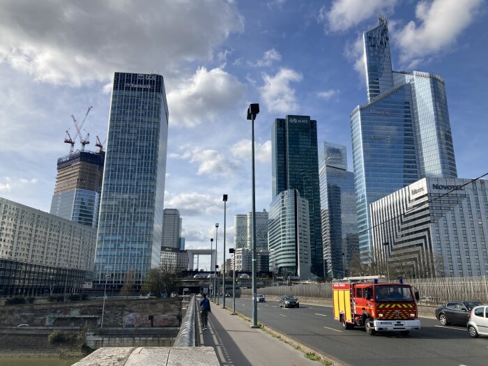 Modernt stadsskapsvy med skyskrapor, en brandbil i rörelse på vägen, och en person som går på trottoaren.