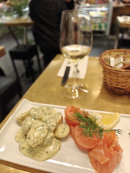 Tallrik med rimmad lax, krämig sås, dill, citronskiva och ett glas vitt vin i bakgrunden.