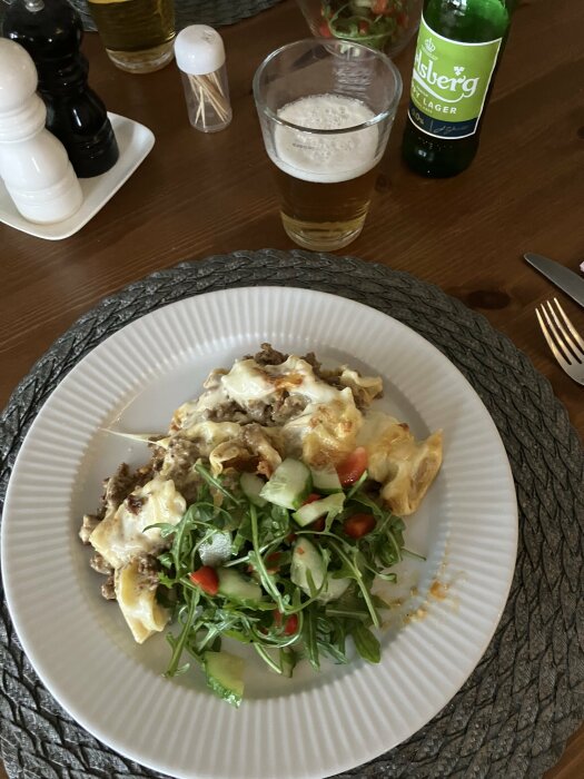 Tallrik med pastagratäng arrabiata, köttfärssås, sallad, kryddor, öl och saltkar på ett bord.