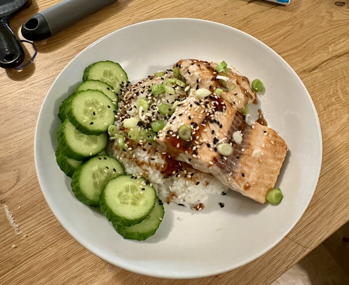 Ris med grillad lax toppad med teriyakisås, sesamfrön och salladslök, serverat med skivad gurka.