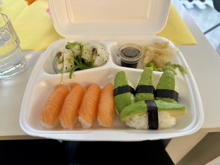 Sushi i take away-förpackning med lax nigiri, avokadomaki, rullar, ingefära, sojasås och wasabi.