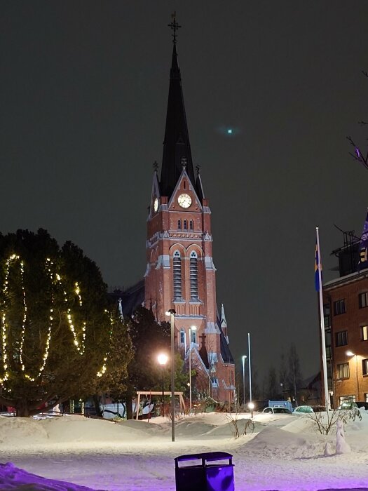 Kyrkobyggnad belyst på kvällen under vintern med snö och dekorerade träd.