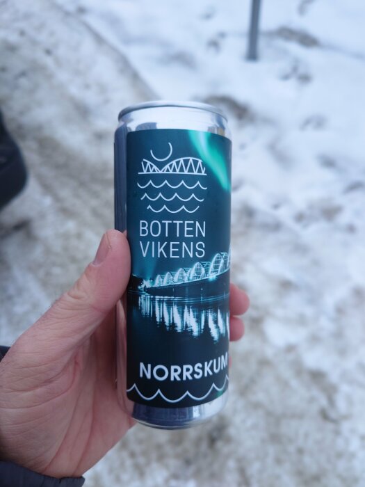 Hand håller en ölburk med texten "Botten Vikens" och "Norrsken" mot bakgrund av snö.