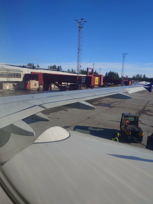 Vy från flygplansfönster med vinge över flygplatsens gater och en markarbetare vid fordon.