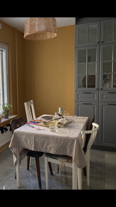 Köksdel med gula väggar och grått skåp, matbord med linneduk och rottinglampa, fönster med växt.