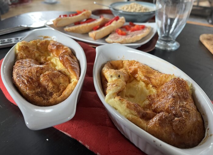 Hembakt ugnsomelett med ost i vita formar på bord med bröd och toppingar i bakgrunden.