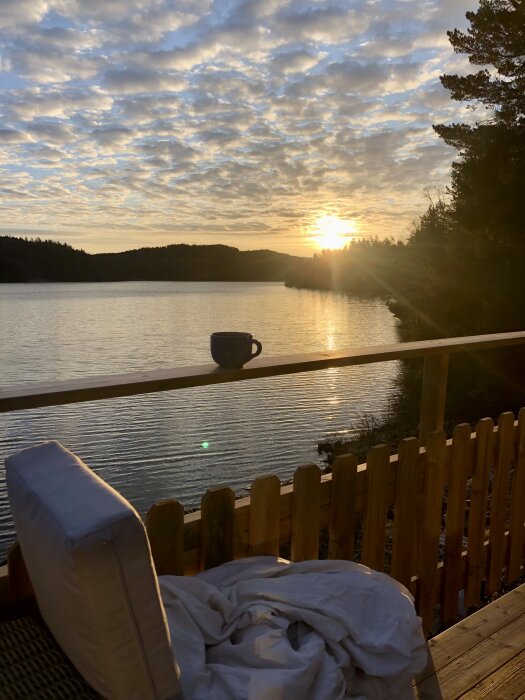 Soluppgång över en sjö sedd från ett trädäck med en kaffekopp på räcket och en soffa med filt.