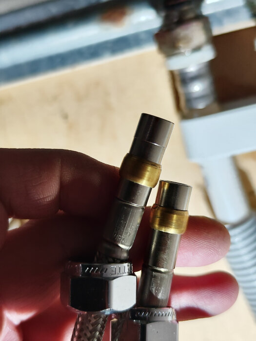 Hand håller två klämringar för rörförbindelse, en med gänga för lin/gängtejp, mot en suddig bakgrund.