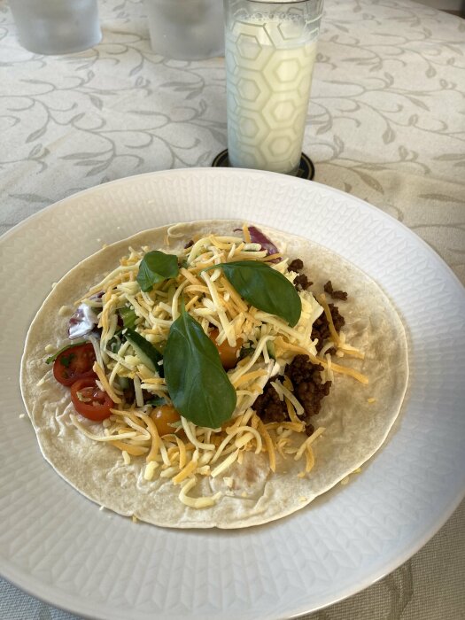 Tortilla med tacokött, ost, sallad och tomater, serverad med ett glas mjölk i bakgrunden.