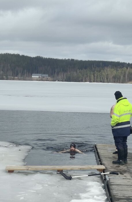 Person i vinterbad håller sig i brygga, observatör i reflexväst står på bryggan, delvis frusen sjö i bakgrunden.