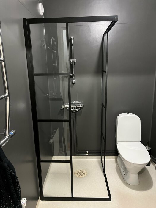 Svart duschhörna med skjutdörrar monterad i ett badrum intill en toalett, väggrör synliga.