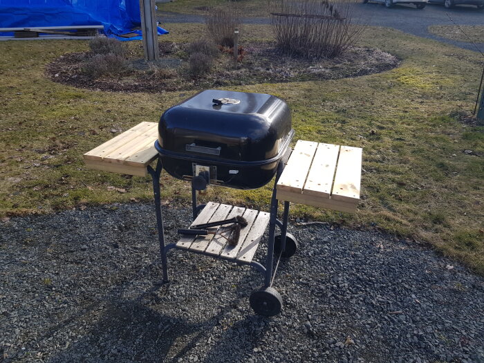 Renoverad grill med nya träsidobord på en grusig yta, omgiven av gräsmatta.