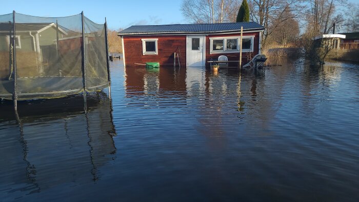 Översvämmat hus och trädgård med möbler delvis under vatten, tydlig skada på fastigheten.