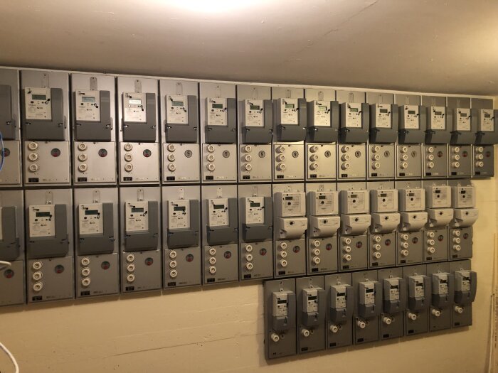 Vägg med rad av elektricitetsmätare och säkringsboxar i ett teknikrum.